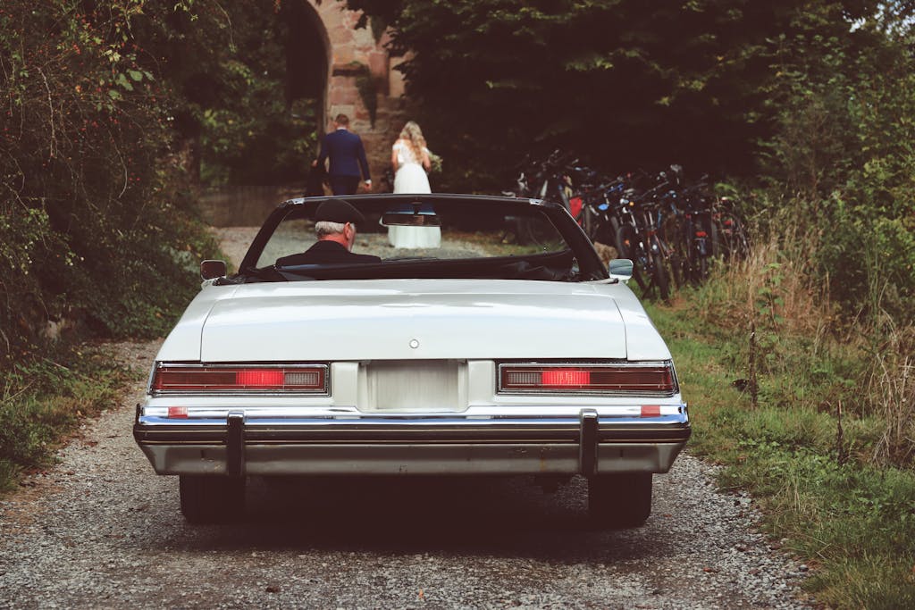 A vintage car parked on a gravel path, with a bride and groom walking ahead.